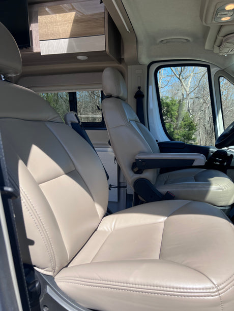 Front cabin of an RV rental with comfortable, light-colored captain's chairs, overhead storage, and a bright outdoor view.