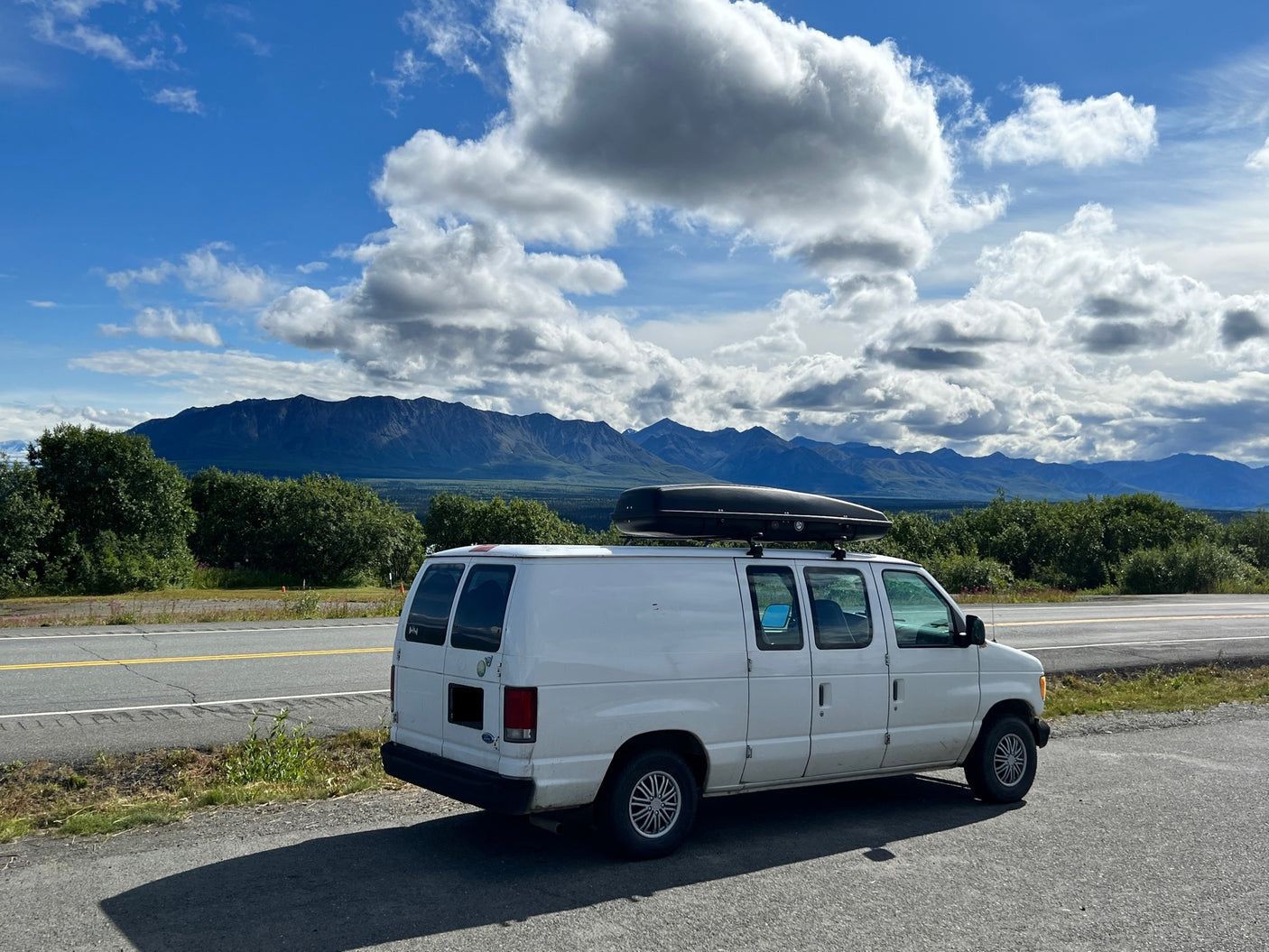 A white van, equipped with a roof box, parked on a scenic road with mountains and clouds. An ideal RV rental for a nature road trip.