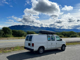 A white van, equipped with a roof box, parked on a scenic road with mountains and clouds. An ideal RV rental for a nature road trip.