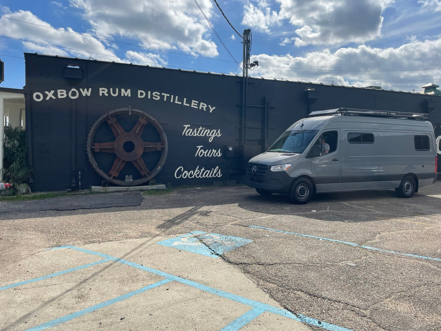 Gray RV rental van parked at Oxbow Rum Distillery, a dark building with 'Tastings Tours Cocktails' and a large gear.