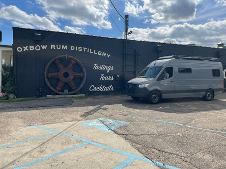Gray RV rental van parked at Oxbow Rum Distillery, a dark building with 'Tastings Tours Cocktails' and a large gear.