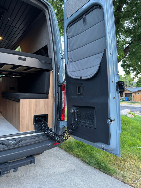 RV rental camper van's open rear reveals a convenient outdoor shower, bed, seating area, and cozy wood interior.