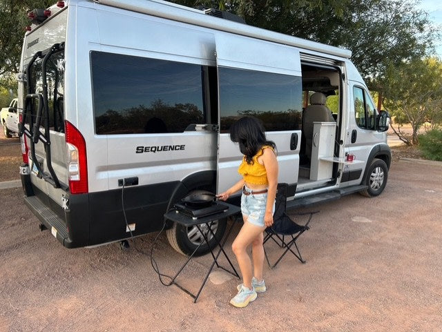 Woman cooks on an outdoor griddle next to a silver RV rental with an open side door and bike rack.