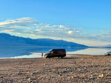 An RV rental parked on rocky shore next to mirror-like water, reflecting blue sky and distant mountains. People paddle on water.