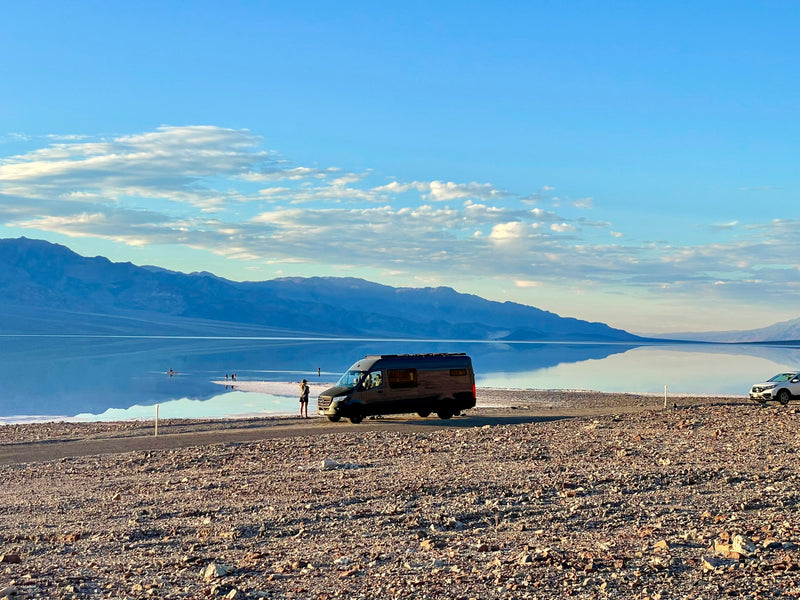An RV rental parked on rocky shore next to mirror-like water, reflecting blue sky and distant mountains. People paddle on water. — photo 7