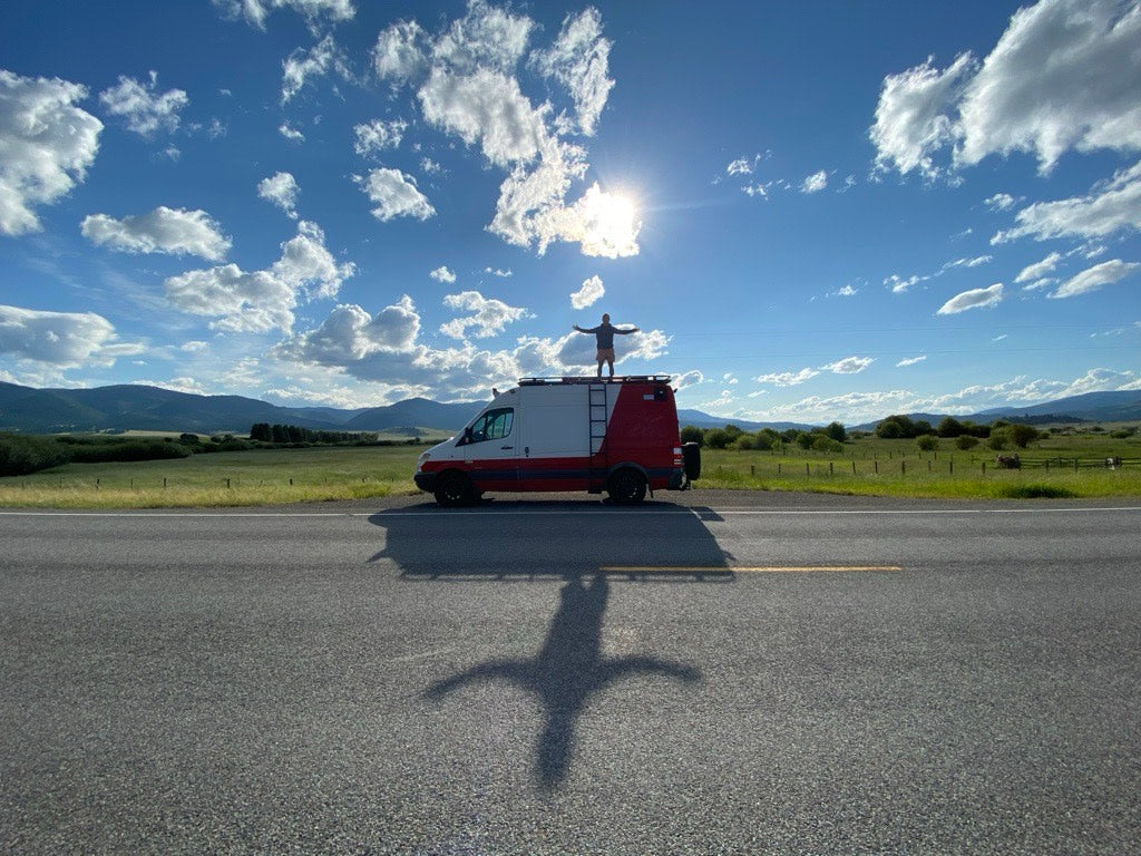 Person standing on an RV rental van, arms outstretched, admiring the scenic landscape. Sun casts a unique shadow.