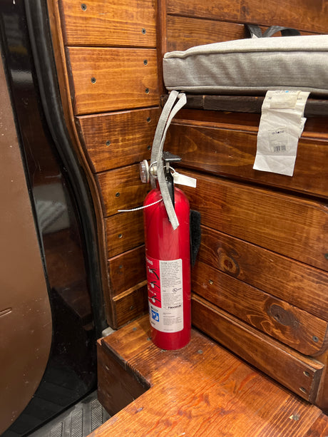 Essential safety gear: a red fire extinguisher secured to the wooden interior of an RV rental, with a grey cushion nearby.