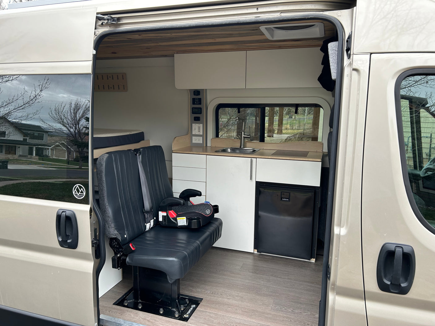 Spacious RV rental camper van interior featuring custom kitchen, bench seating with child booster, and rear bed.