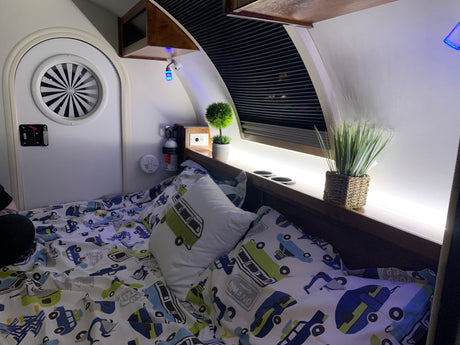 Cozy RV rental sleeping area: bed with car-themed bedding, circular window, blue accent & LED lighting, safety gear.