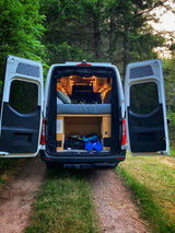 Rear doors of an RV rental open to reveal a comfortable bed and storage inside, set in a peaceful wooded area.
