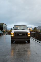 This white van, with headlights on, sits on a wet surface. Could be part of an rv rental fleet.