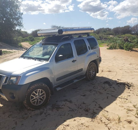 Silver Nissan Xterra SUV with a rooftop tent on a sandy road, ready for adventure camping. Explore with this RV rental alternative.