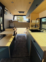 Stylish RV rental interior: bed, modern kitchen with sink, light wood accents, & dark cabinetry along a plush rug runner.