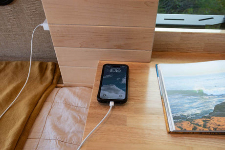 RV rental interior: Phone charging on a light wood surface next to an open book showing ocean waves, ideal for travel.