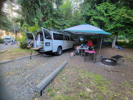 Camper van (RV rental) set up at a wooded campsite with a canopy and picnic area. Person cooking, kids nearby.