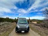 Front view of a gray Winnebago campervan RV rental on a dirt path, surrounded by hills and cloudy blue sky.