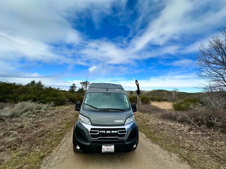 Front view of a gray Winnebago campervan RV rental on a dirt path, surrounded by hills and cloudy blue sky.