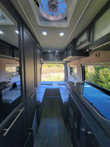 Spacious RV rental interior with kitchenette, lounge/bed, and overhead storage, looking out to nature.