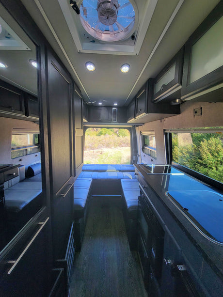 Spacious RV rental interior with kitchenette, lounge/bed, and overhead storage, looking out to nature.