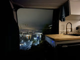 Night view from an RV rental: a kitchen with gold faucet overlooks city lights, a fenceline, and water. Mobile living awaits!