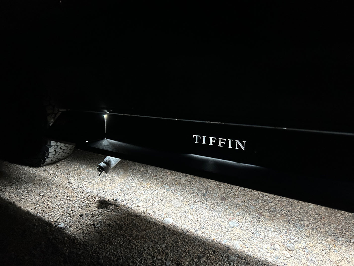 Night shot of an RV rental undercarriage light illuminating gravel, showing 'TIFFIN' side branding and tire. Ready for travel.