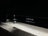 Night shot of an RV rental undercarriage light illuminating gravel, showing 'TIFFIN' side branding and tire. Ready for travel.