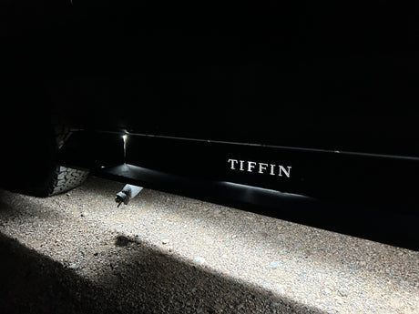 Night shot of an RV rental undercarriage light illuminating gravel, showing 'TIFFIN' side branding and tire. Ready for travel.