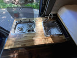 Inside an RV rental: a compact kitchen featuring a two-burner stove, stainless steel sink, and wood countertop.