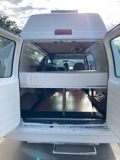 RV rental camper van interior. Features a convertible bed/bench, built-in storage, and visible front cab, ready for travel.
