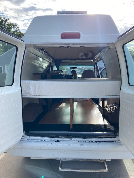 RV rental camper van interior. Features a convertible bed/bench, built-in storage, and visible front cab, ready for travel.