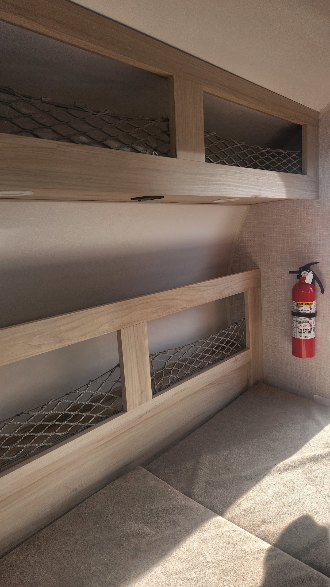 RV rental interior: light wood storage with netting, a padded area, and a red fire extinguisher mounted on a textured wall.