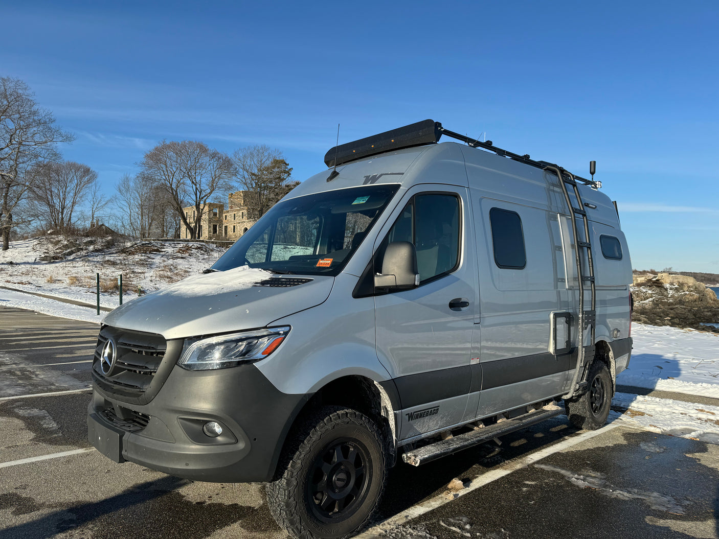 Grey Mercedes-Benz rv rental van with roof rack, ladder, and snow, parked near old stone ruins.