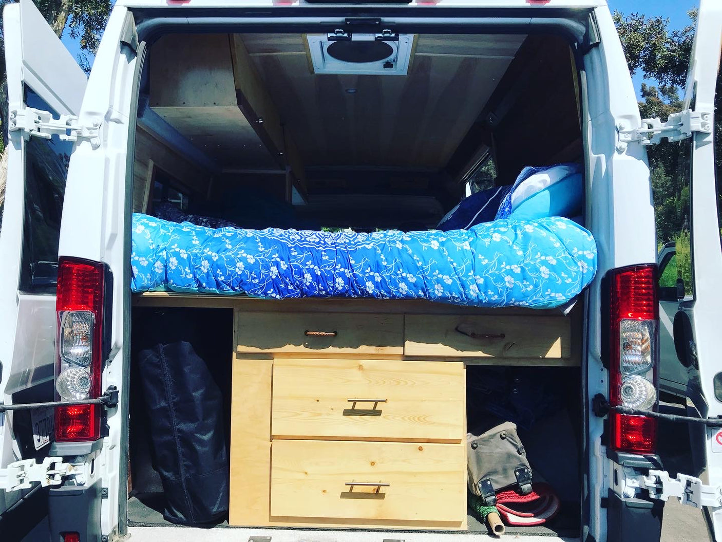 Back of an RV rental van, revealing a cozy blue-patterned bed above custom wooden storage. Ideal for road trips & camping.