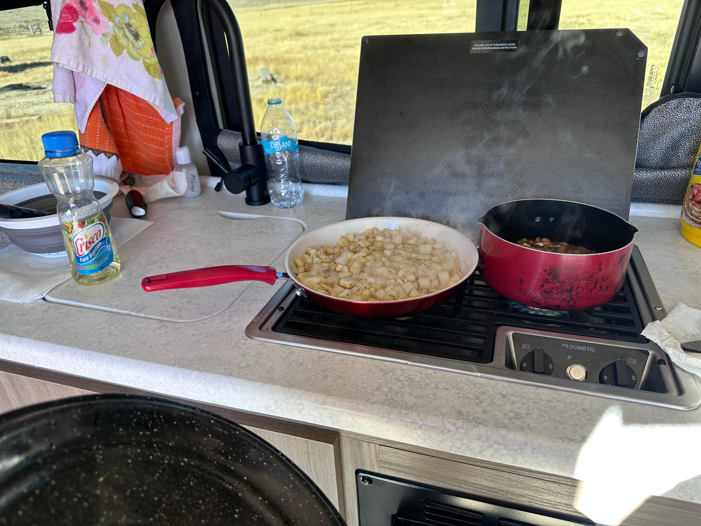 Preparing a warm meal in an RV rental kitchen: potatoes and beans cooking on a Dometic gas stove, with oil and water ready.