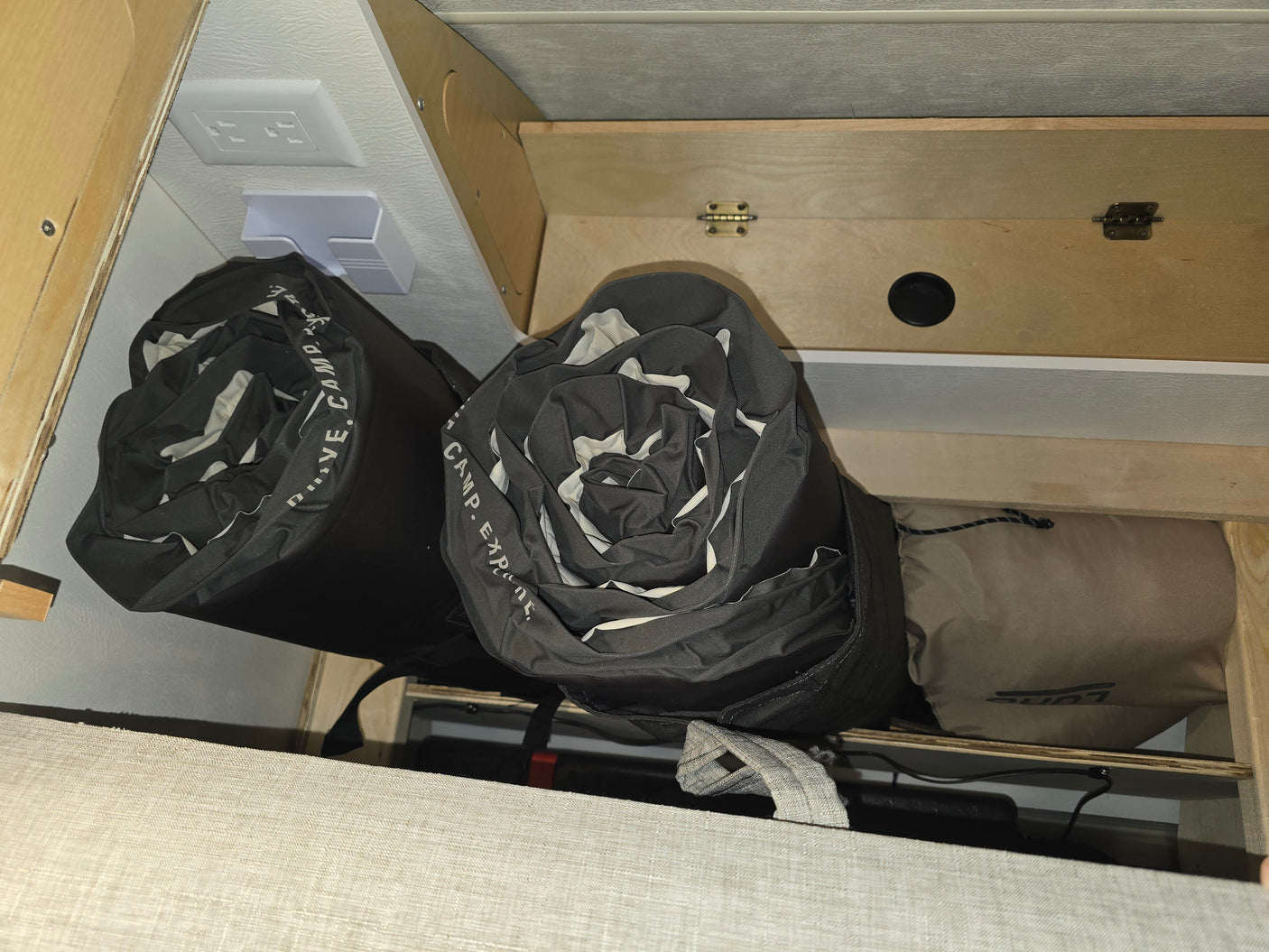 RV rental storage: Rolled sleeping bags, a power outlet, and camping gear are neatly stowed under a bed or bench.
