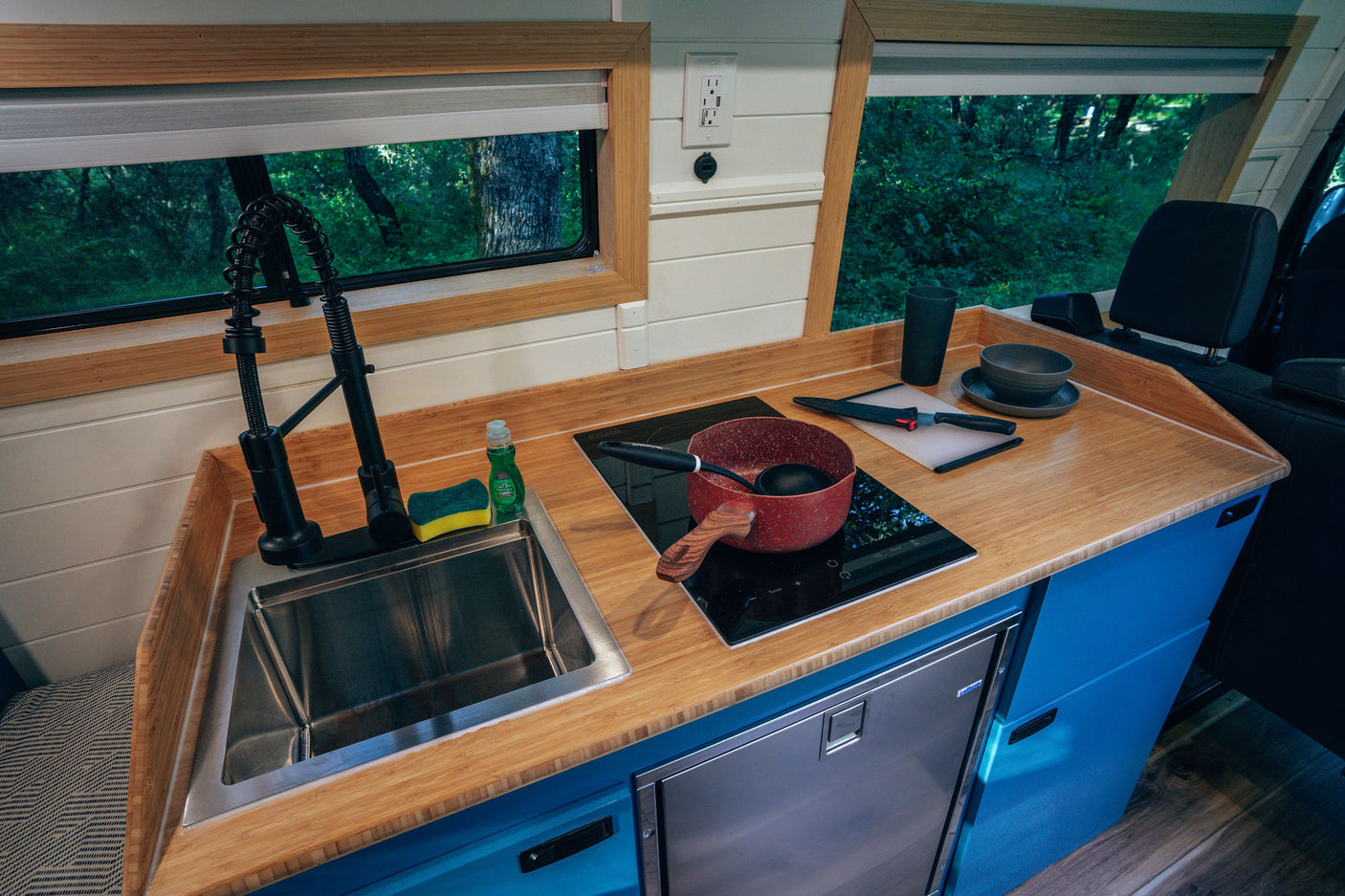 Compact RV rental kitchen with bamboo counters, sink, induction cooktop, and fridge, offering forest views.
