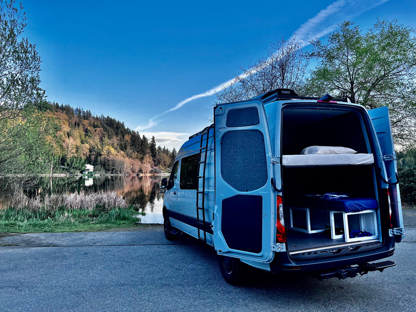 A light blue RV rental camper van with open back doors revealing bunk beds, parked by a serene lake and forested hills. — photo 12