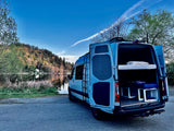 A light blue RV rental camper van with open back doors revealing bunk beds, parked by a serene lake and forested hills.