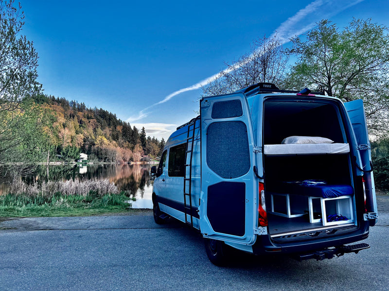 A light blue RV rental camper van with open back doors revealing bunk beds, parked by a serene lake and forested hills. — photo 12
