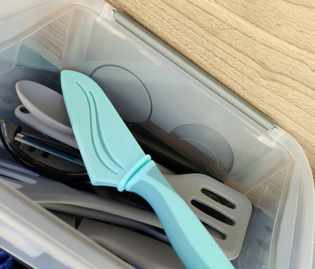 RV rental kitchen: clear bin of organized utensils, including a blue pie server & grey spatula for your travels.