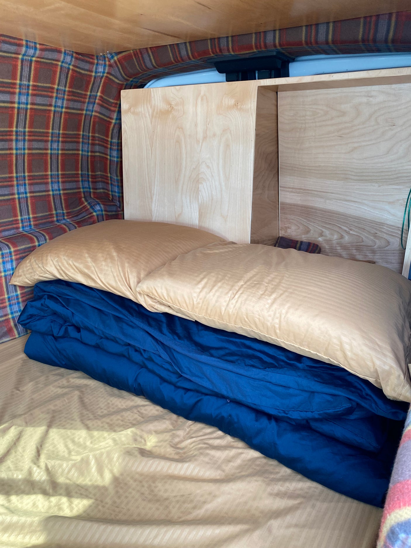 Compact RV rental sleeping area: bed with blue blanket, beige pillow, plaid walls, & wood paneling.