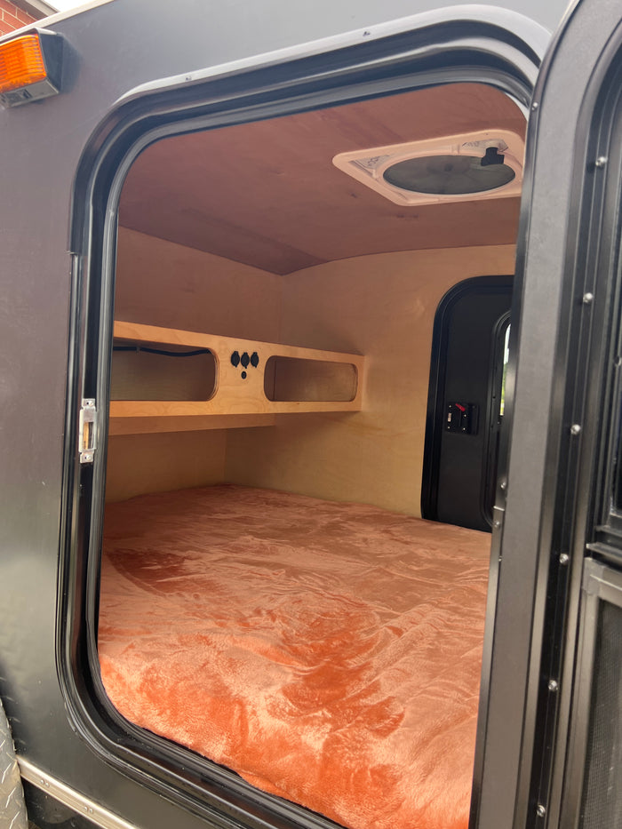 Welcoming RV rental interior: orange bed, wood-paneled walls/ceiling, shelf with outlets, and open door revealing the exterior.