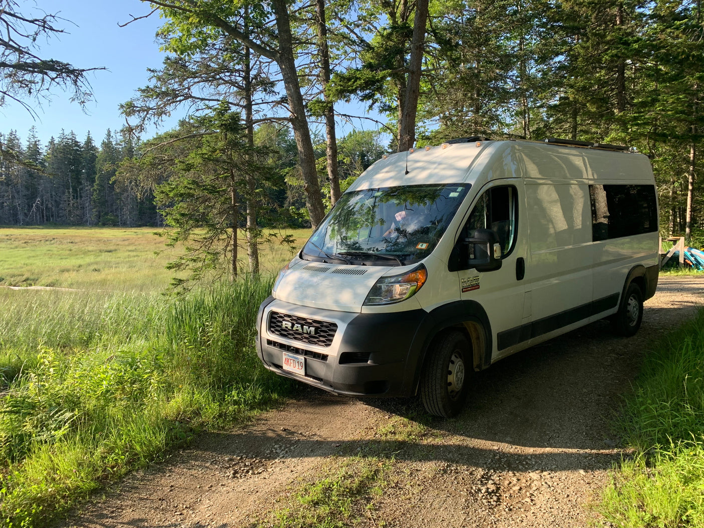 RV rental van parked on a dirt road next to a lush green marsh and towering forest under a clear sky.
