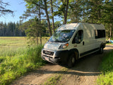 RV rental van parked on a dirt road next to a lush green marsh and towering forest under a clear sky.