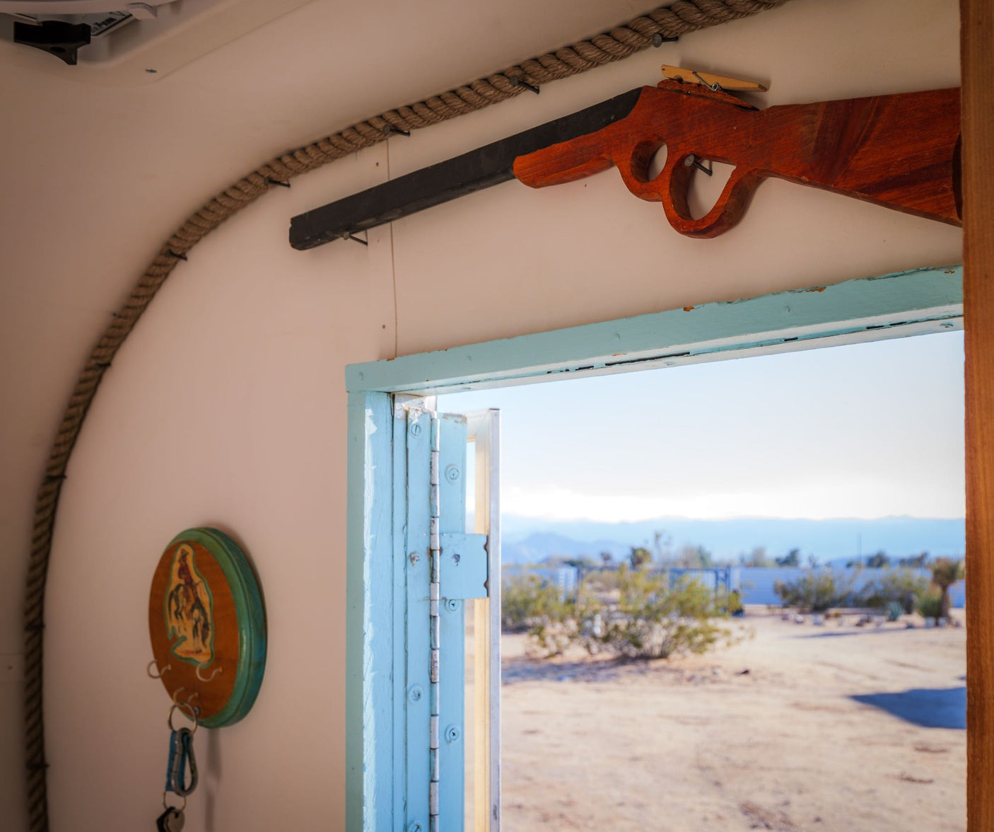 RV rental interior featuring a decorative wooden rifle above a light blue door, with rope trim, key hooks, and a desert view.
