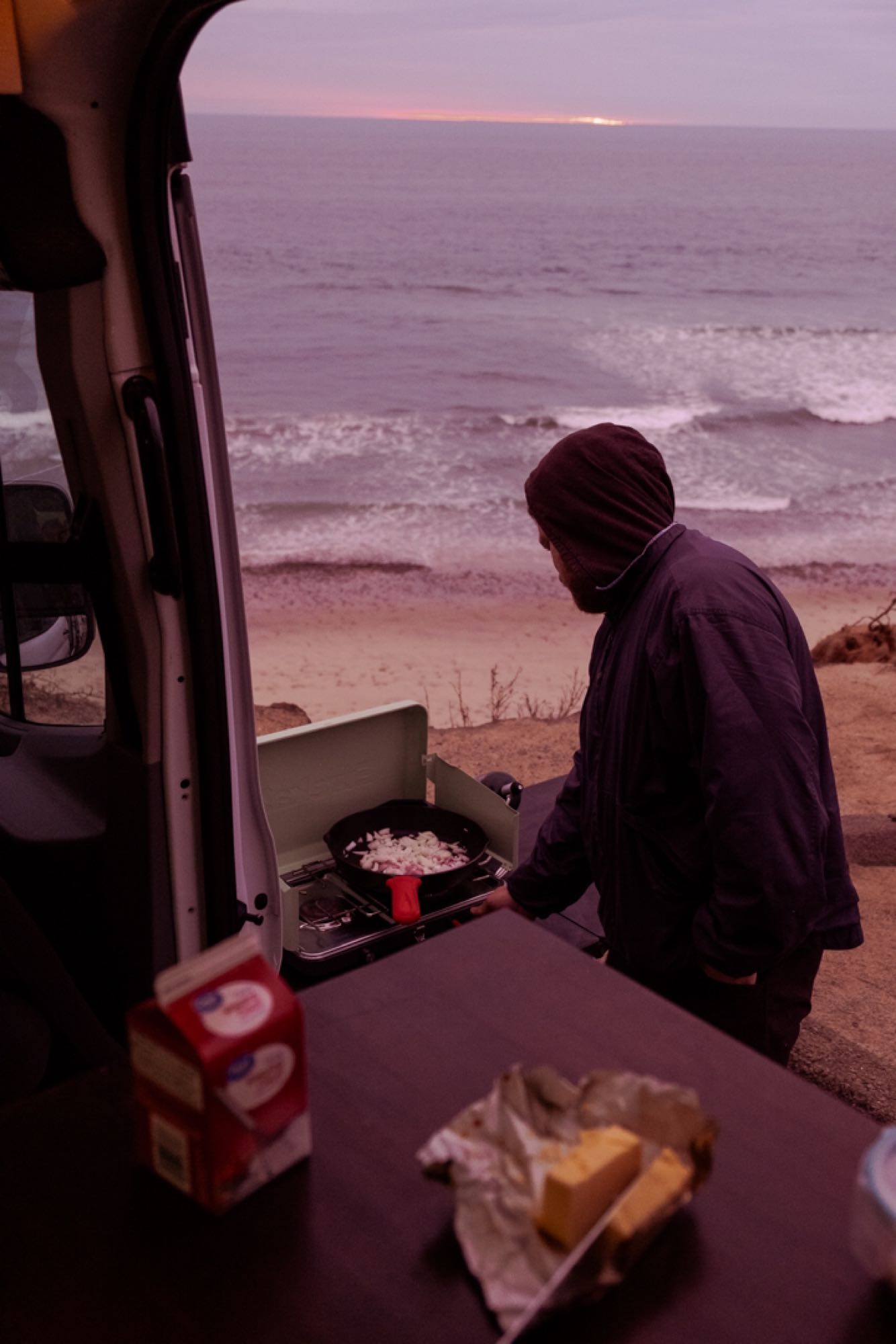 A person cooks onions on a portable stove by the ocean at sunrise from their rv rental, with waves in the background.