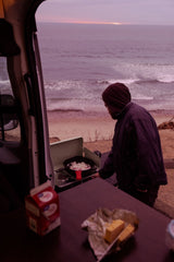 A person cooks onions on a portable stove by the ocean at sunrise from their rv rental, with waves in the background.