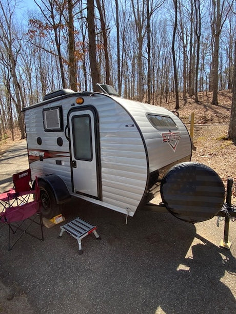 Silver teardrop rv rental travel trailer parked in a sunny wooded campground. Features a spare tire with an American flag cover.