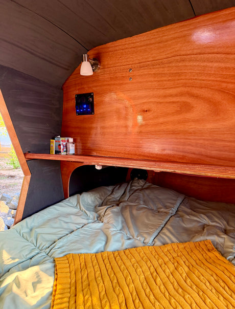 An RV rental's comfy interior with a bed, wooden walls, lighting, and power controls showing 13.9V.