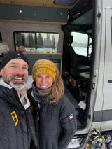 A smiling couple bundled in winter gear stands in front of their open rv rental camper van during a snowy adventure. Snowshoes are ready!